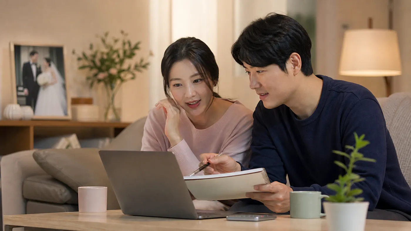 신혼부부가 집에서 노트북과 노트를 보며 생활비와 저축 계획을 대화하는 모습